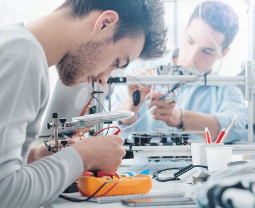 Engineering students working in the lab, a student is using a voltage and current tester, another student in the background is using a 3D printer